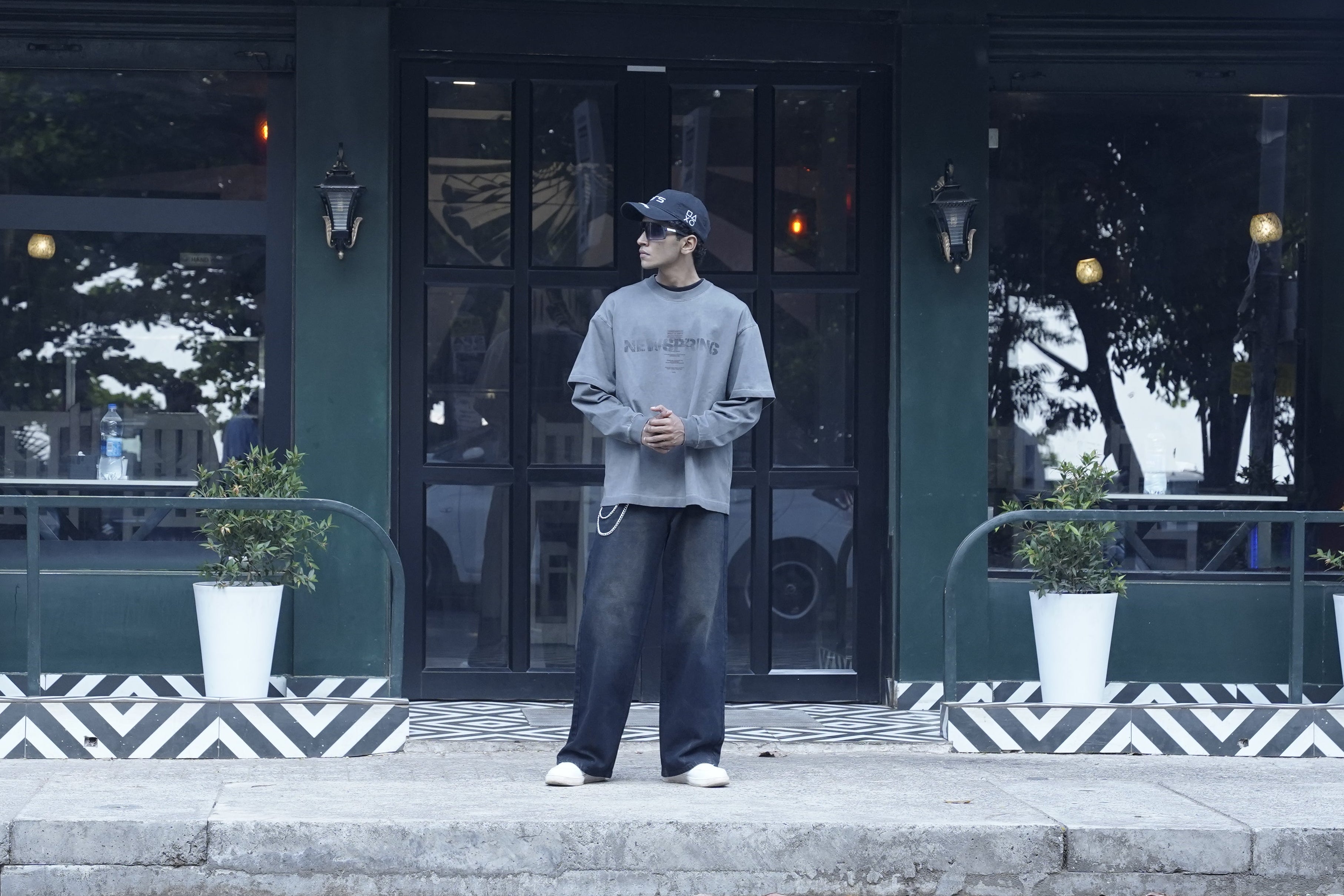 Person wearing a gray sweatshirt and dark pants standing on a street with a building in the background
